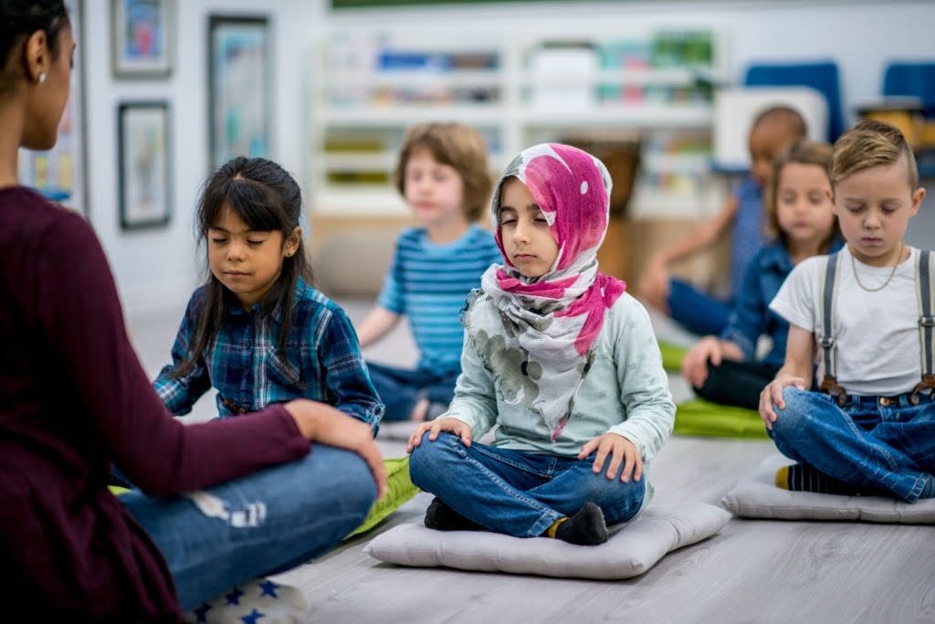 A multi-ethnic group of young school children are indoors in their classroom. They are sitting on pillows and doing yoga together. They are sitting with their hands in their lap